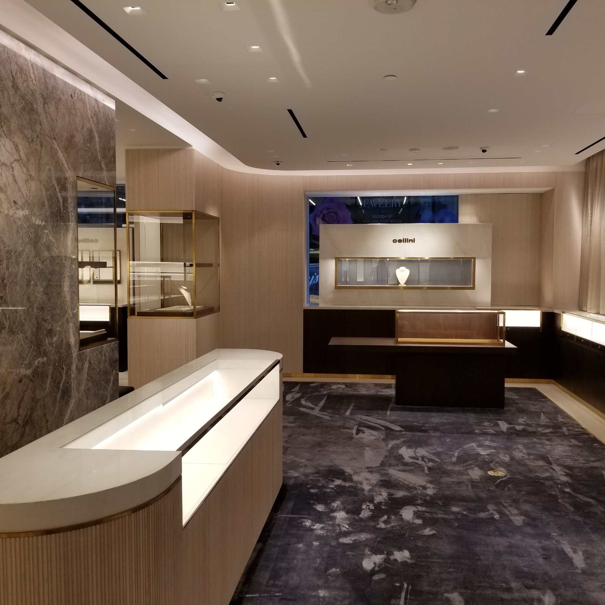Wnętrze luksusowego sklepu jubilerskiego z marmurowymi ścianami, oświetlonymi witrynami i ciemną podłogą z abstrakcyjnym wzorem.