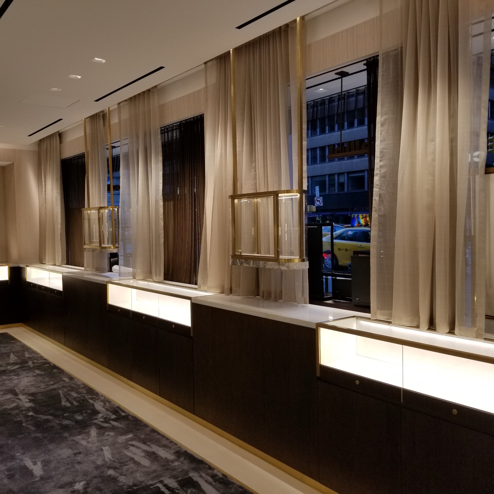 Wnętrze sklepu z ciemnymi drewnianymi szafkami ekspozycyjnymi podświetlonymi od wewnątrz, marmurową podłogą i oknami z jasnymi zasłonami, przez które widać budynki i taksówkę na zewnątrz.