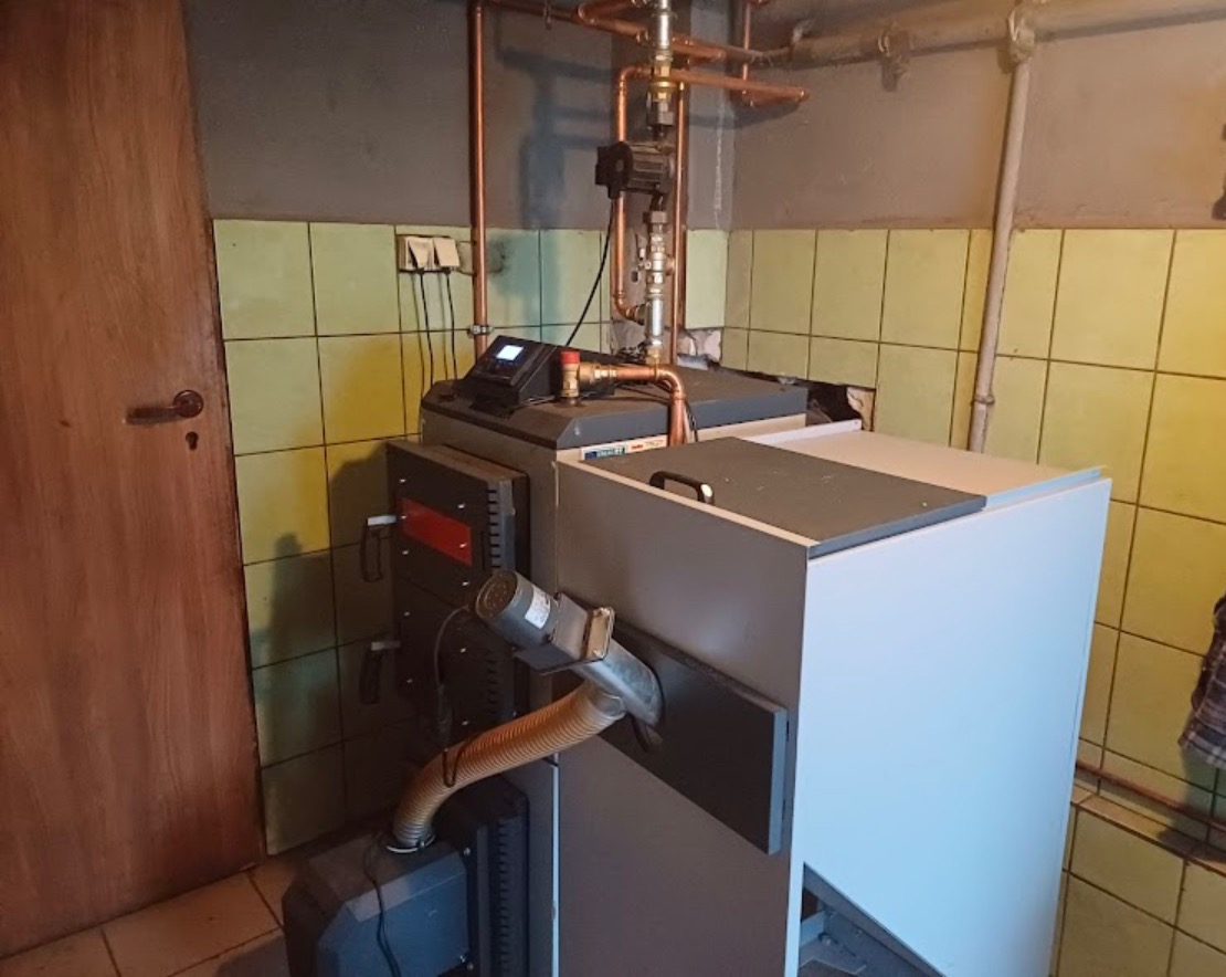 Nowoczesny piec na paliwo stałe z elektronicznym panelem sterowania, rurami miedzianymi i wentylatorem, zainstalowany w kotłowni z wykafelkowanymi ścianami.