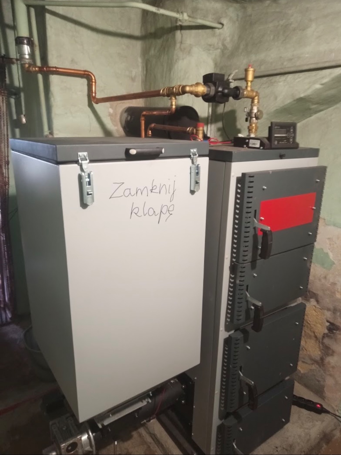 Nowoczesny piec na paliwo stałe z napisem 'Zamknij klapę' na białej obudowie, widoczne miedziane rury instalacji grzewczej i elementy sterujące.