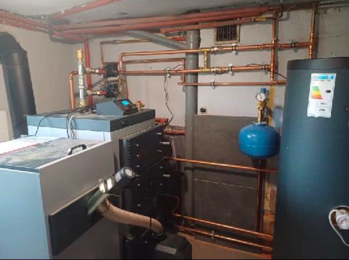 Kompleksowa instalacja centralnego ogrzewania z kotłem na paliwo stałe, widoczne miedziane rury, zbiornik wyrównawczy oraz panel sterowania, zainstalowane w piwnicy.