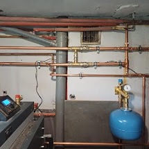 Złożona instalacja centralnego ogrzewania z rur miedzianych, widoczny fragment kotła z panelem sterowania, szary komin wentylacyjny i niebieski zbiornik wyrównawczy, całość w piwnicy z surowym...