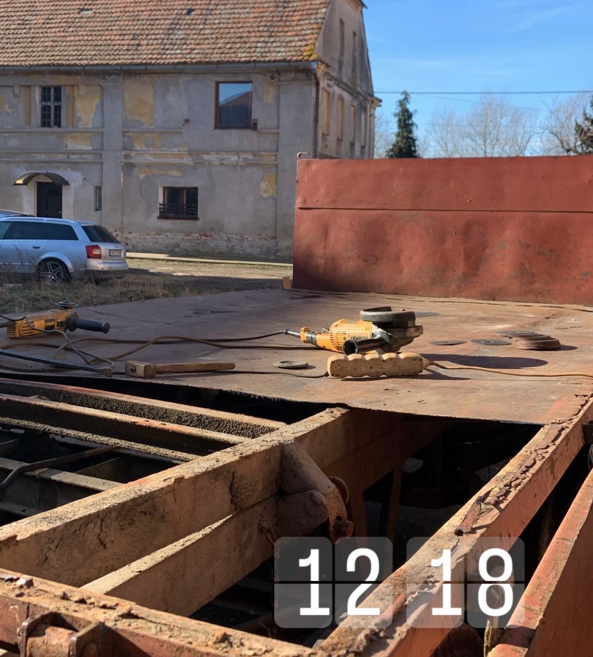 Rusty metalowa konstrukcja z narzędziami do obróbki metalu, w tle budynek z odpadającym tynkiem i zaparkowany samochód.