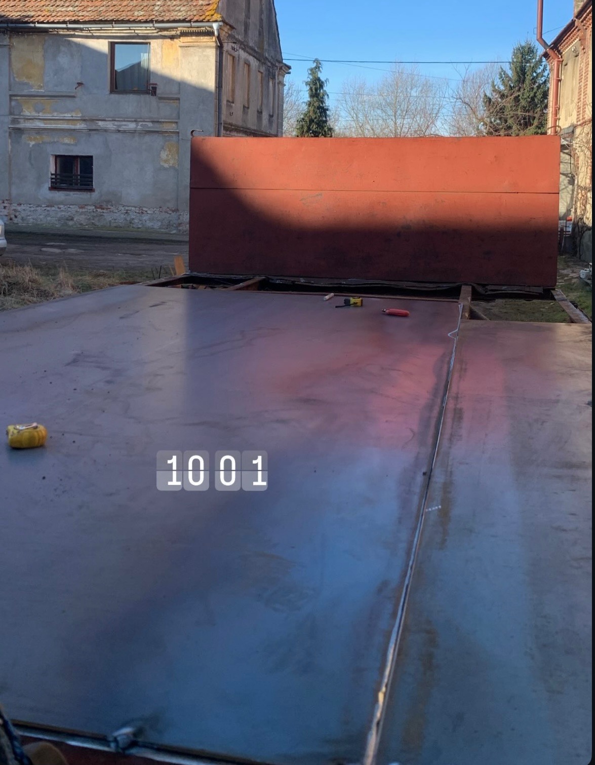 Duża, metalowa konstrukcja w trakcie budowy na zewnątrz, widoczne narzędzia pomiarowe i spoiny, w tle budynek mieszkalny.