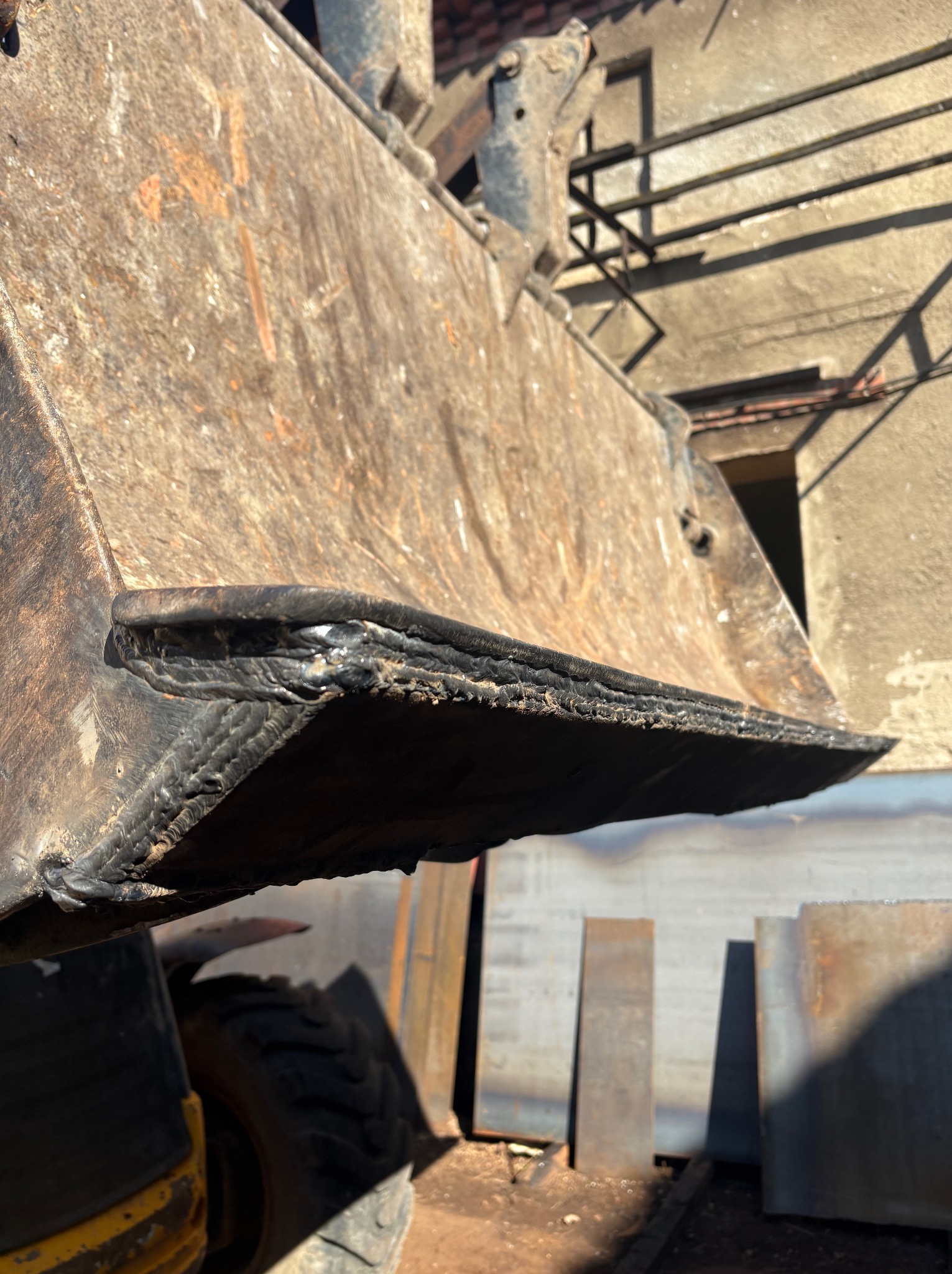 Przód łyżki koparki, widoczne świeże spawy wzmacniające krawędź, z bliska, w tle blachy i fragment budynku.