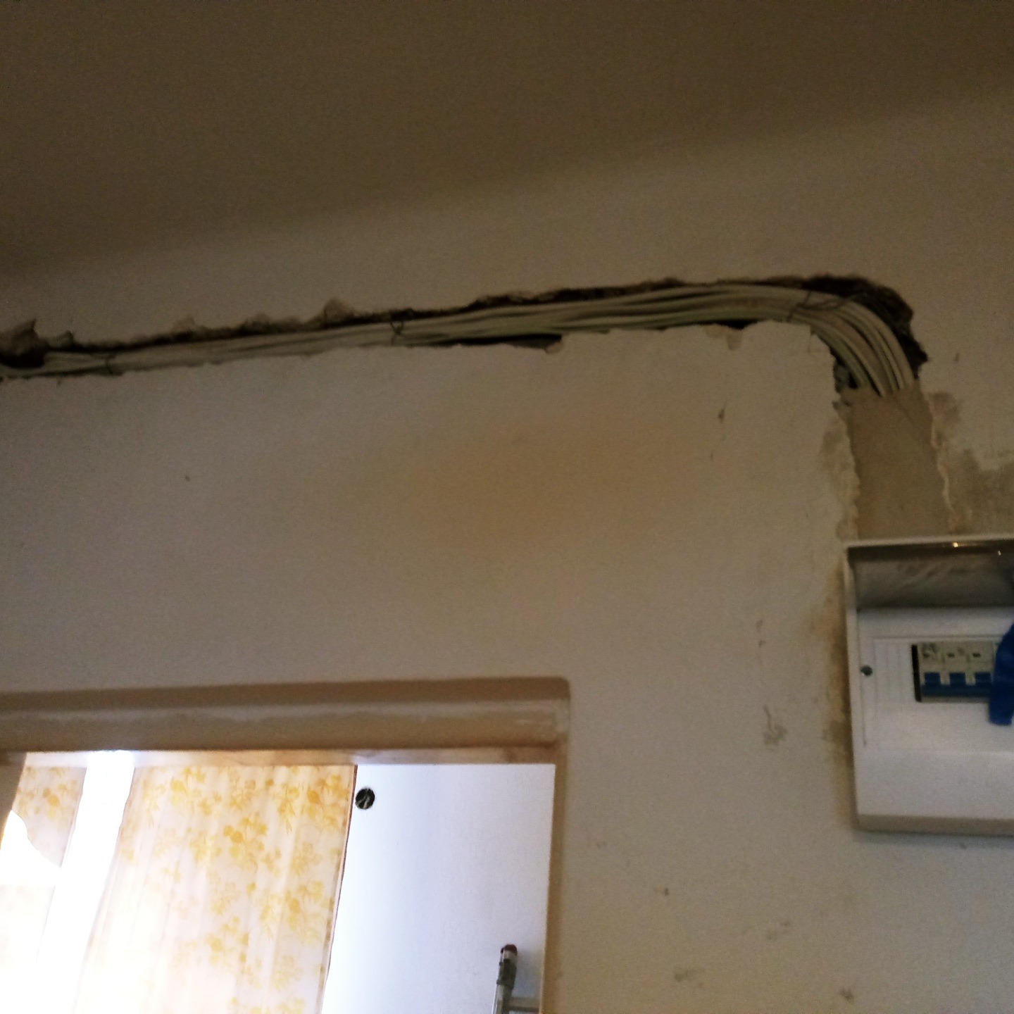 Wykute w ścianie kable elektryczne prowadzone pod sufitem, widoczna skrzynka z bezpiecznikami i fragment ościeżnicy drzwiowej.