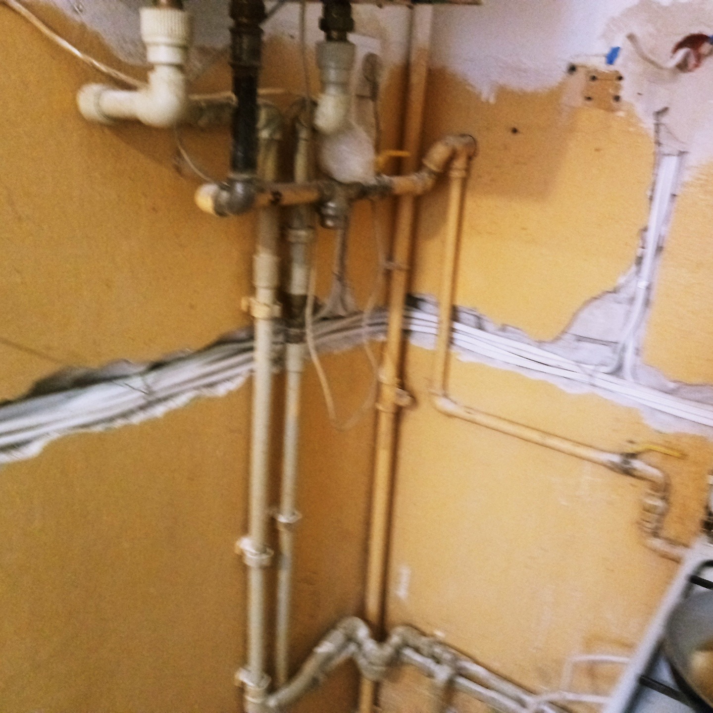 Fragment ściany w trakcie wymiany instalacji elektrycznej i wodnej, widoczne przewody elektryczne, rury wodne i gazowe, uszkodzony tynk i fragment kuchenki.