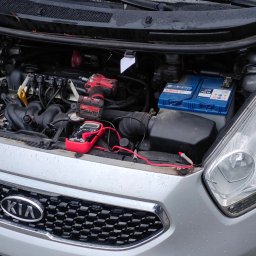 Pomoc drogowa Mobilny serwis HOL-MOBIL - Otwarta maska srebrnego samochodu marki Kia, widoczny akumulator Bosch, multimetr z czerwonymi przewodami oraz zakrętak udarowy Milwaukee, sugerujące prace diagnostyczne lub naprawcze.