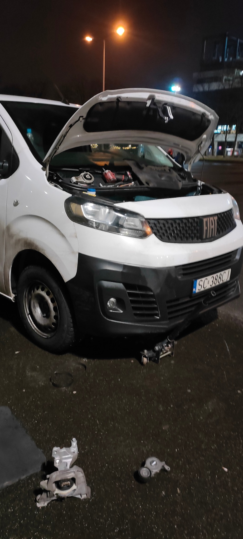 Biały samochód dostawczy marki Fiat z otwartą maską, pod którą widoczne są narzędzia i części; na ziemi leżą zdemontowane elementy, a pod przednim zderzakiem znajduje się podnośnik.