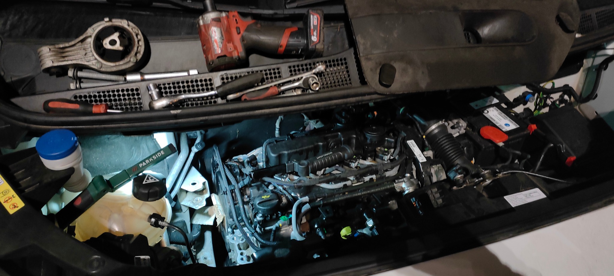 Silnik samochodowy w trakcie naprawy, widoczne narzędzia, akumulator, zbiornik płynu chłodniczego i lampa warsztatowa Parkside.