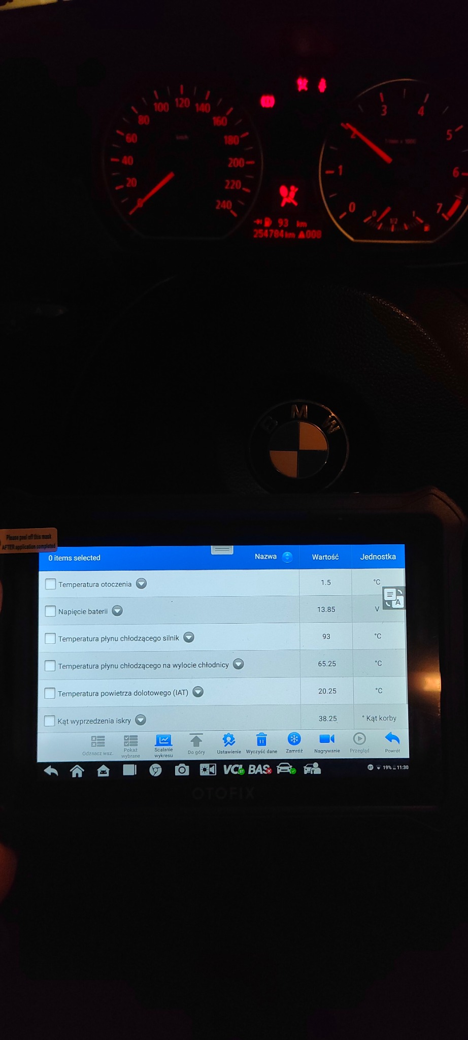 Ekran urządzenia diagnostycznego z odczytami temperatury płynu chłodzącego, napięcia baterii i kąta wyprzedzenia zapłonu w samochodzie BMW, widoczne zegary deski rozdzielczej i logo marki.