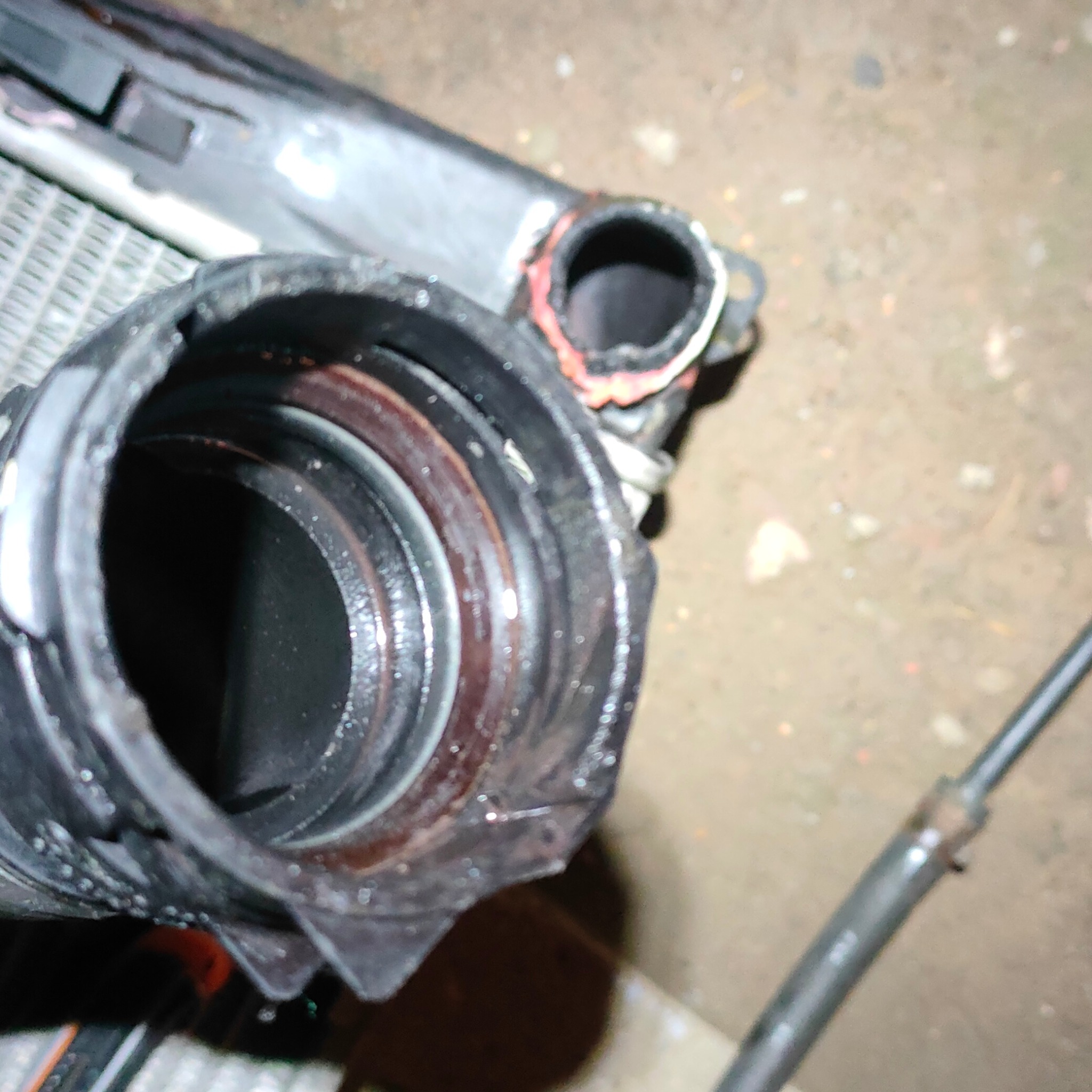 Zbliżenie na otwarty wylot chłodnicy samochodowej z widocznymi uszczelkami i resztkami płynu chłodniczego, na tle fragmentu kratownicy chłodnicy i przewodu gumowego.