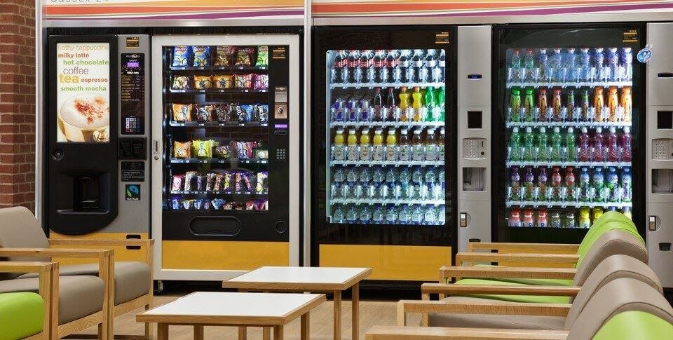 Automaty sprzedające napoje, słodycze i kawę w pomieszczeniu z miejscami do siedzenia.