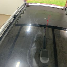 Filip Padziński - Widok z góry na brudny dach samochodu z zamontowanym bagażnikiem dachowym, widoczny mechanizm otwierania bramy garażowej i fragment ogrodu przez otwartą bramę.