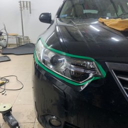 Filip Padziński - Czarny samochód z zieloną taśmą zabezpieczającą wokół reflektora, polerka elektryczna leży na podłodze w warsztacie, przygotowanie do polerowania lakieru.