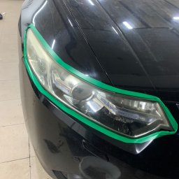 Filip Padziński - Przygotowanie reflektora do polerowania, zabezpieczonego zieloną taśmą malarską na czarnym samochodzie w Urzędowie.