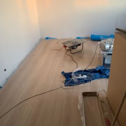 Filip Padziński - Proces układania paneli podłogowych: widoczna piła do cięcia paneli, rozłożone panele, listwy przypodłogowe zabezpieczone folią, oraz przedłużacz na niebieskiej folii ochronnej.
