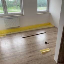 Filip Padziński - Układanie paneli podłogowych w jasnym pokoju, widoczne folia ochronna, panel w trakcie montażu oraz miara zwinięta przy ścianie.