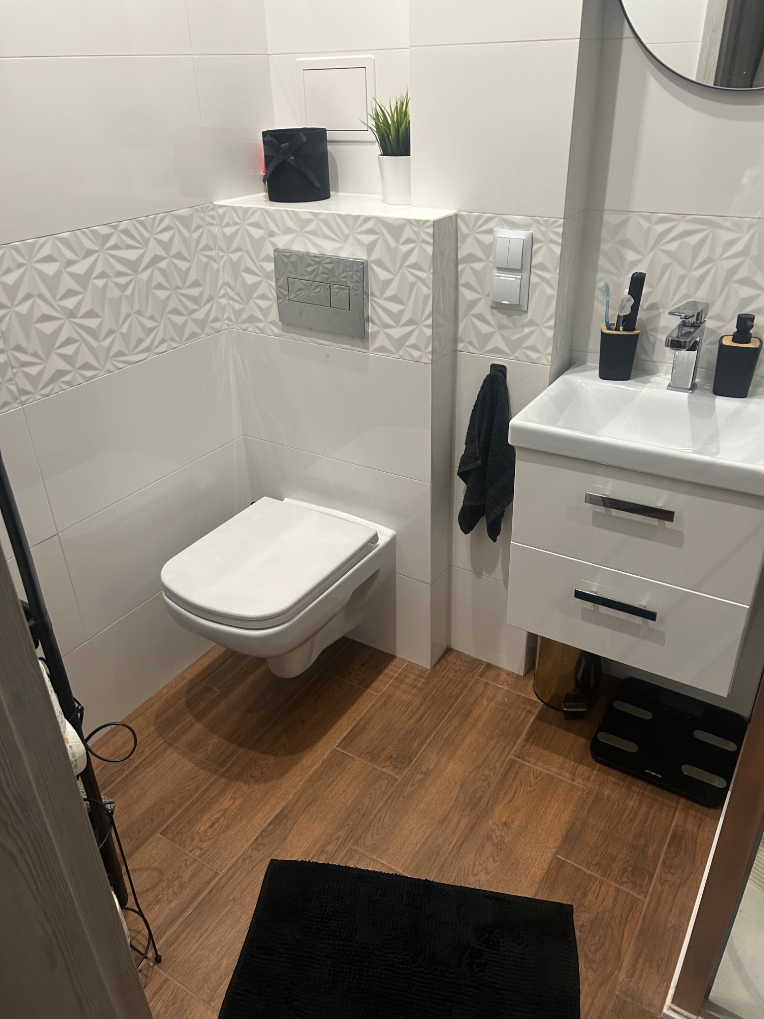 Nowoczesna, jasna łazienka z drewnopodobną podłogą, wiszącą toaletą, umywalką z szafką i dekoracyjnymi płytkami ściennymi z trójwymiarowym wzorem.