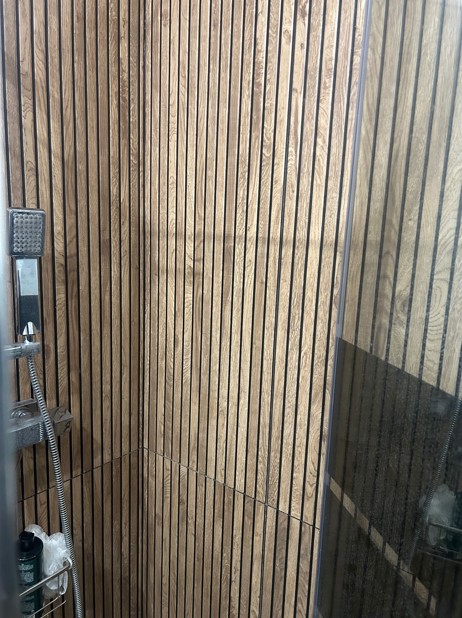 Kabina prysznicowa z wykończeniem ścian z drewnianych paneli pionowych, widoczna słuchawka prysznicowa i półka z kosmetykami.