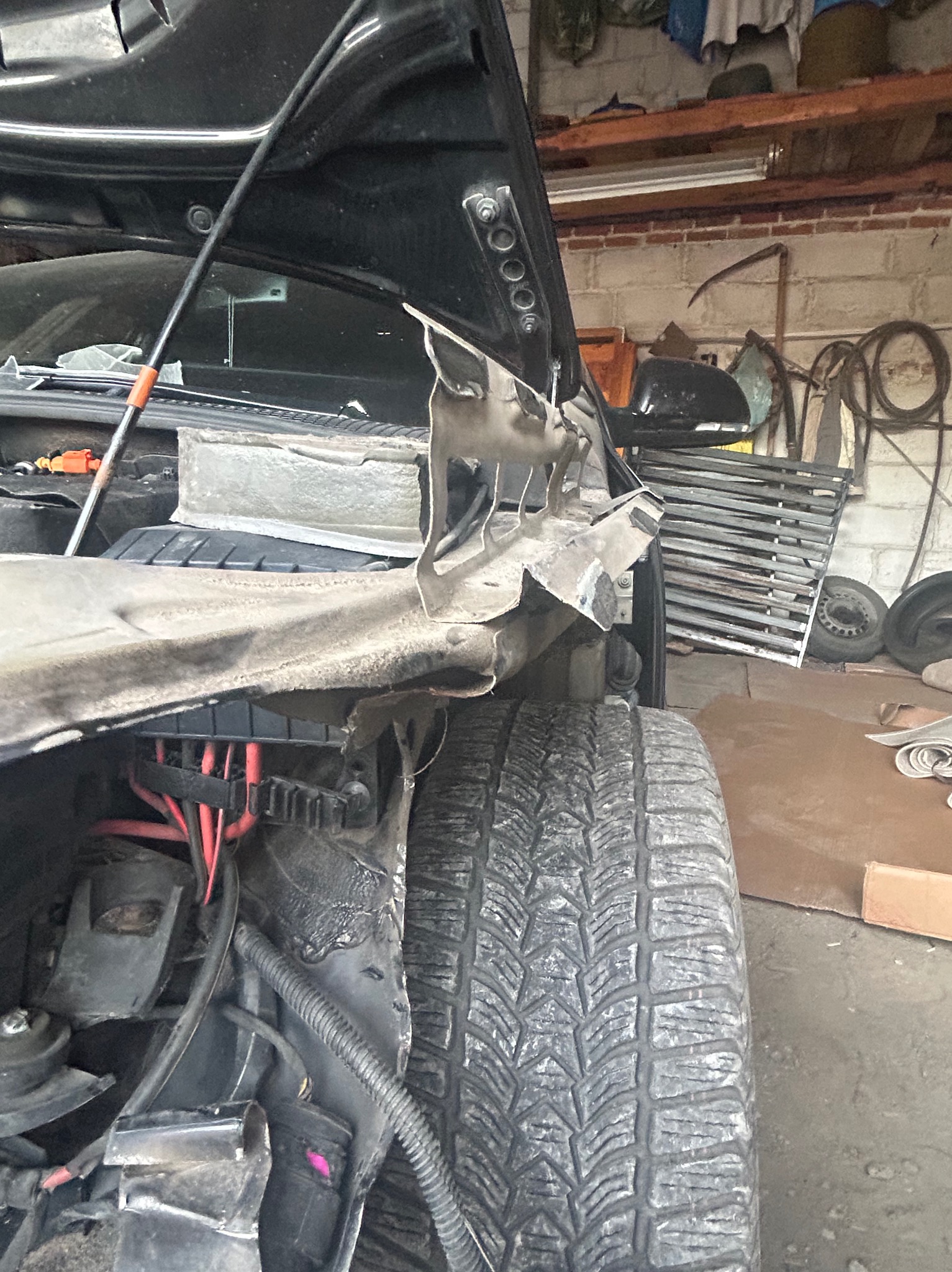 Uszkodzony przód czarnego samochodu z otwartą maską, widoczne zniszczone elementy karoserii i opona z wyraźnym bieżnikiem w warsztacie.