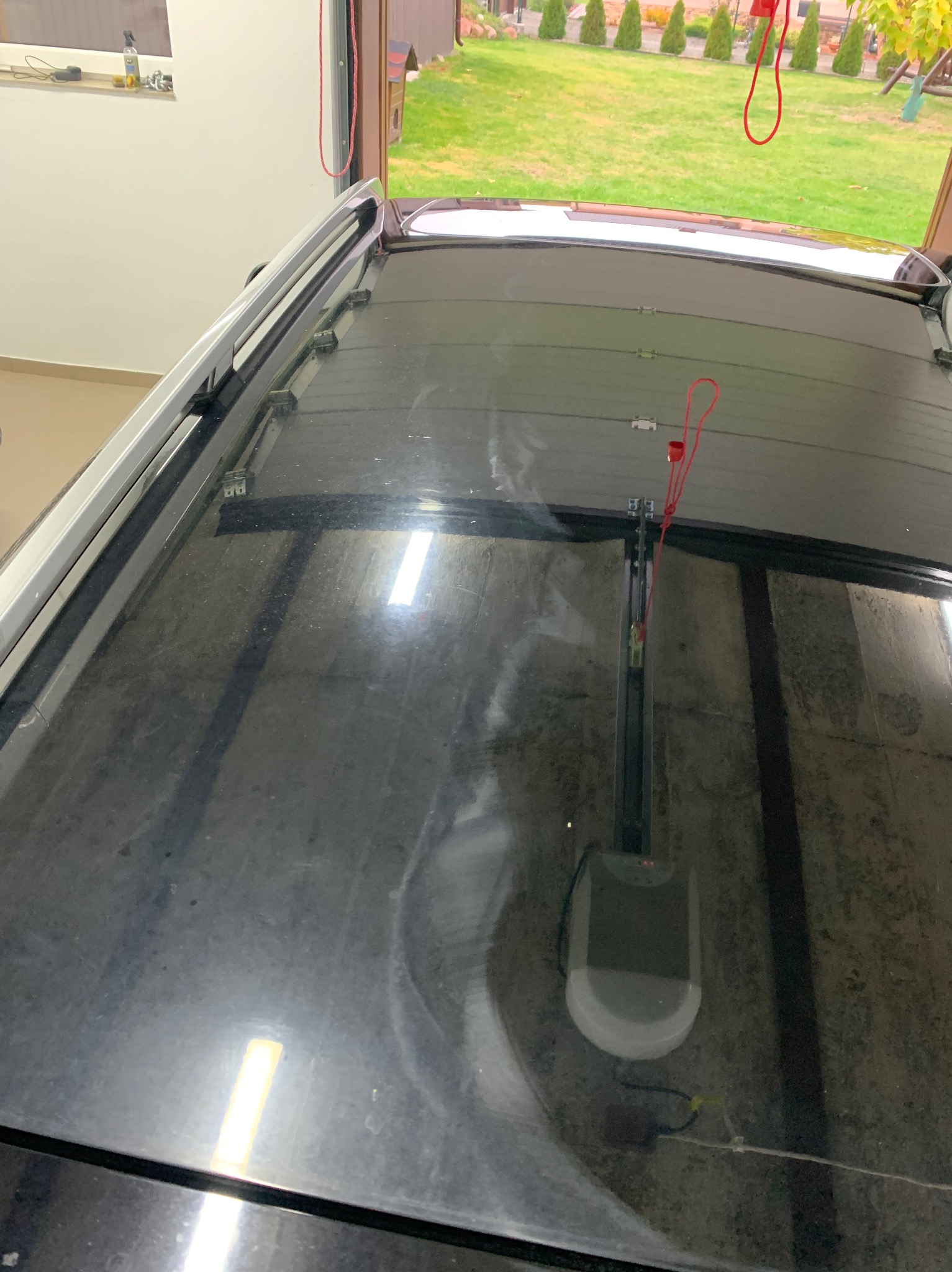 Widok z góry na brudny dach samochodu z zamontowanym bagażnikiem dachowym, widoczny mechanizm otwierania bramy garażowej i fragment ogrodu przez otwartą bramę.