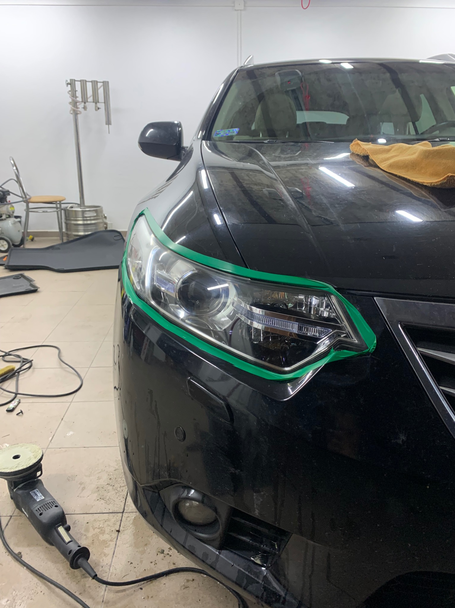 Czarny samochód z zieloną taśmą zabezpieczającą wokół reflektora, polerka elektryczna leży na podłodze w warsztacie, przygotowanie do polerowania lakieru.