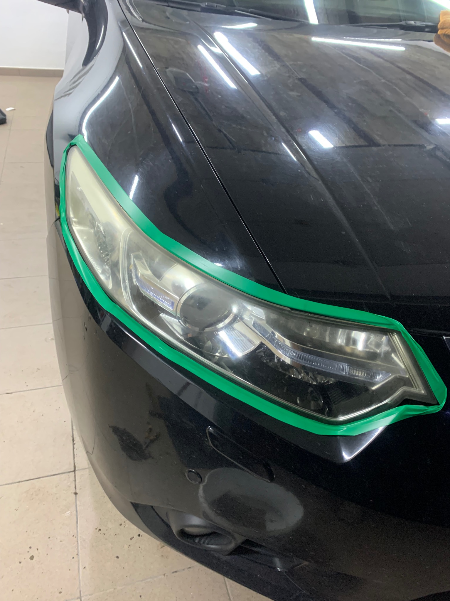 Przygotowanie reflektora do polerowania, zabezpieczonego zieloną taśmą malarską na czarnym samochodzie w Urzędowie.