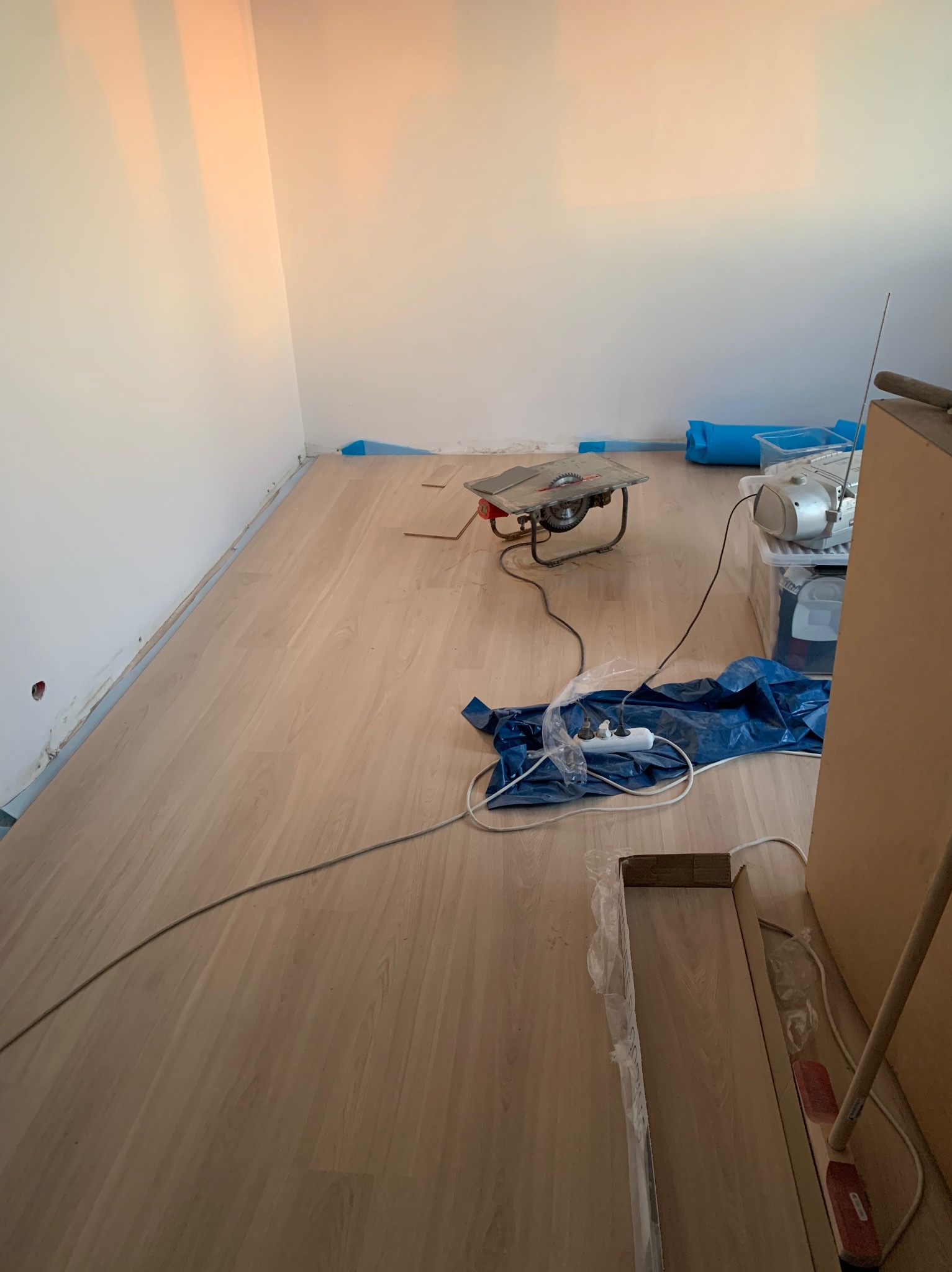 Proces układania paneli podłogowych: widoczna piła do cięcia paneli, rozłożone panele, listwy przypodłogowe zabezpieczone folią, oraz przedłużacz na niebieskiej folii ochronnej.