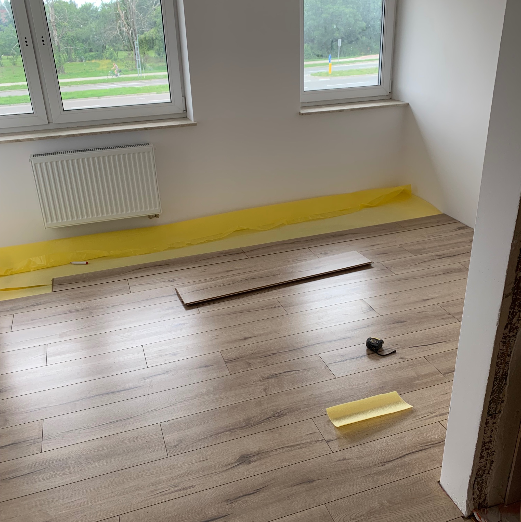 Układanie paneli podłogowych w jasnym pokoju, widoczne folia ochronna, panel w trakcie montażu oraz miara zwinięta przy ścianie.