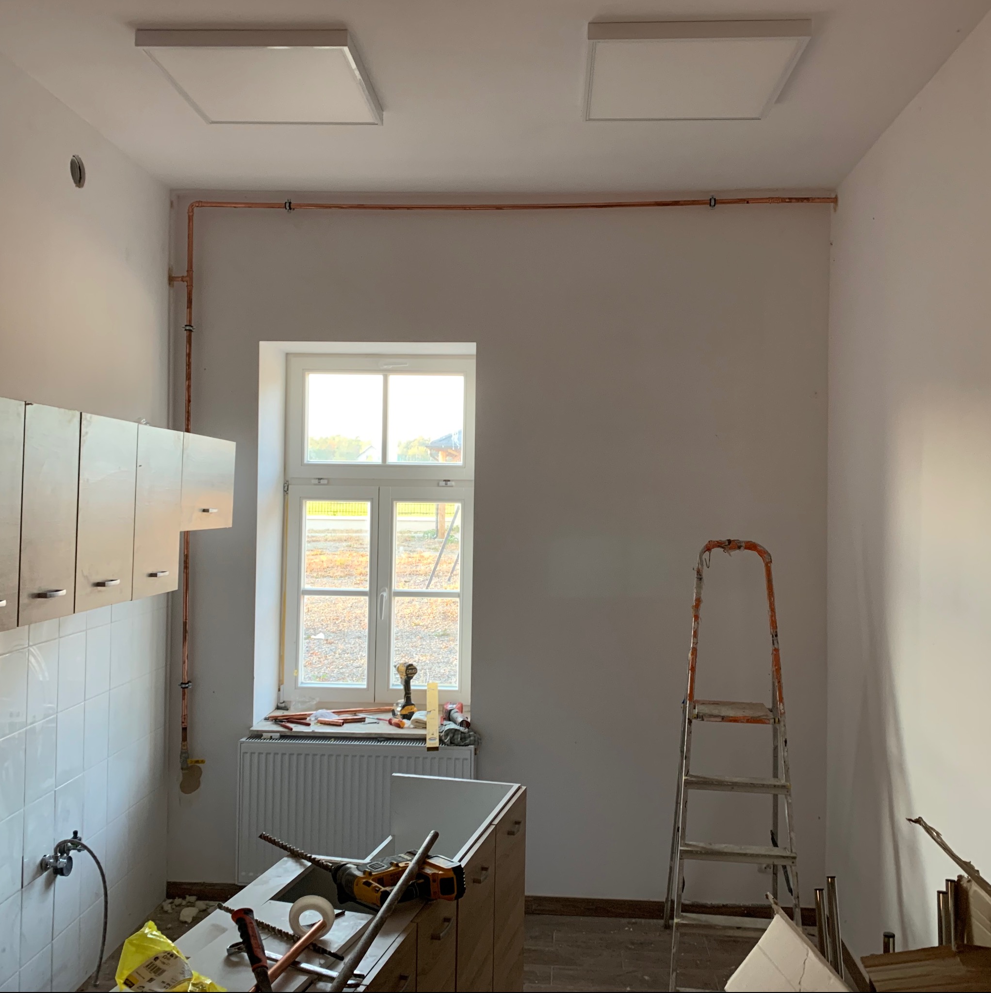 Wnętrze w trakcie remontu: widoczne miedziane rury instalacji wodnej, drabina, częściowo zamontowane szafki kuchenne, narzędzia na blacie oraz dwa panele LED na suficie.