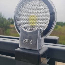 LEPRI SPÓŁKA Z OGRANICZONĄ ODPOWIEDZIALNOŚCIĄ - Lampa ostrzegawcza KEY Automation zamontowana na czarnym słupku bramy, z widocznym logo firmy i tłem z zielenią i pochmurnym niebem.