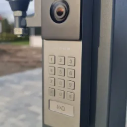 Szary wideofon Vidos z klawiaturą numeryczną i kamerą, zamontowany na metalowym słupku bramy wjazdowej, widok z bliska.