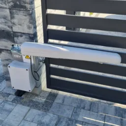 Szara brama posesyjna z zamontowanym napędem automatycznym KEY Automation, widoczna skrzynka sterująca z kablem i ostrzeżeniem o napięciu, kostka brukowa na podjeździe.