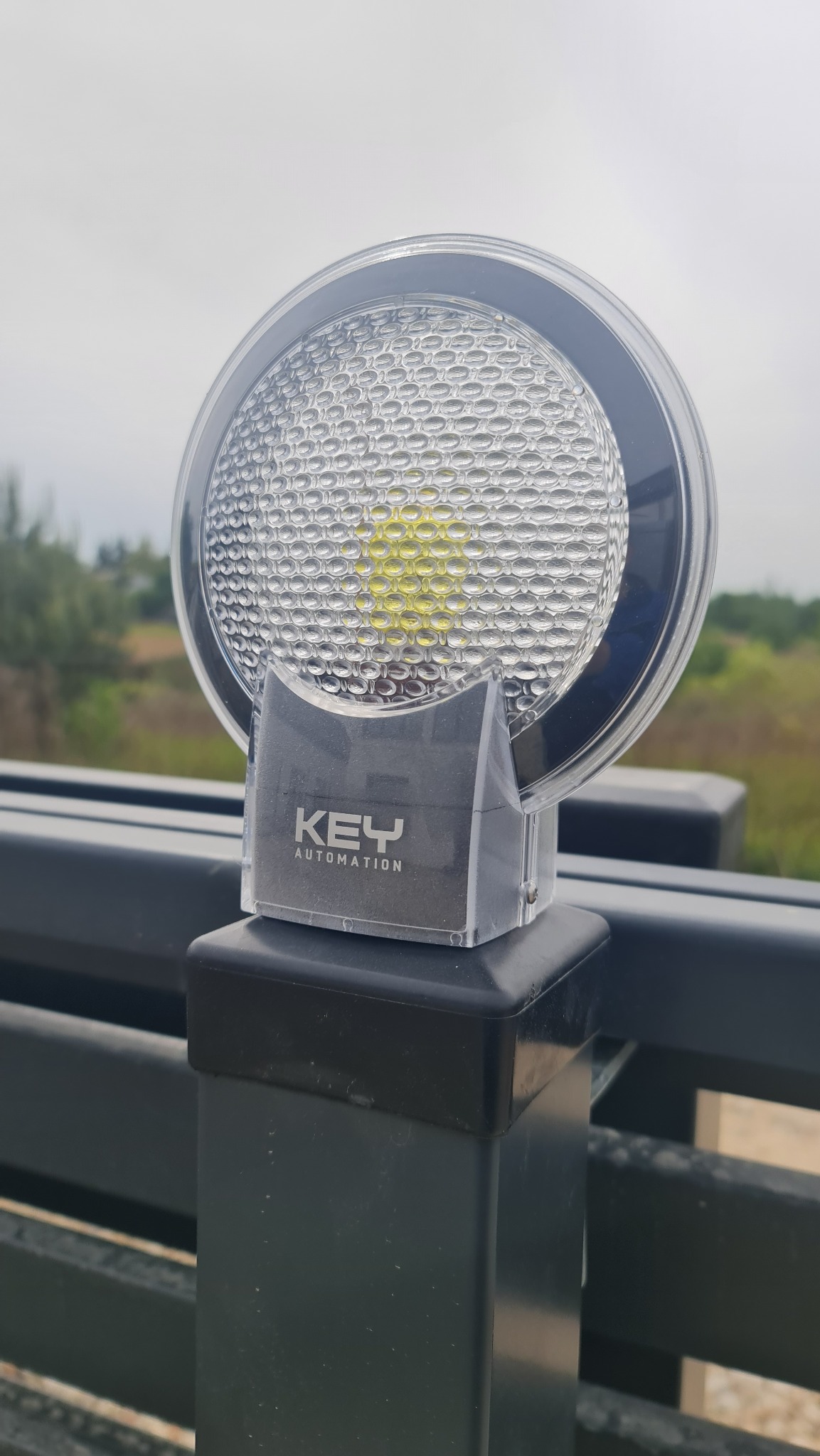 Lampa ostrzegawcza KEY Automation zamontowana na czarnym słupku bramy, z widocznym logo firmy i tłem z zielenią i pochmurnym niebem.