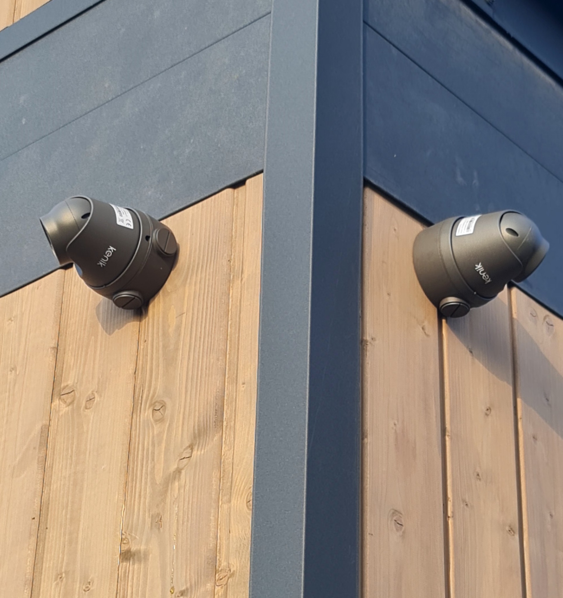 Dwa szare reflektory zewnętrzne marki Kenik zamontowane na drewnianej elewacji budynku z szarymi panelami, oświetlające narożnik budynku.