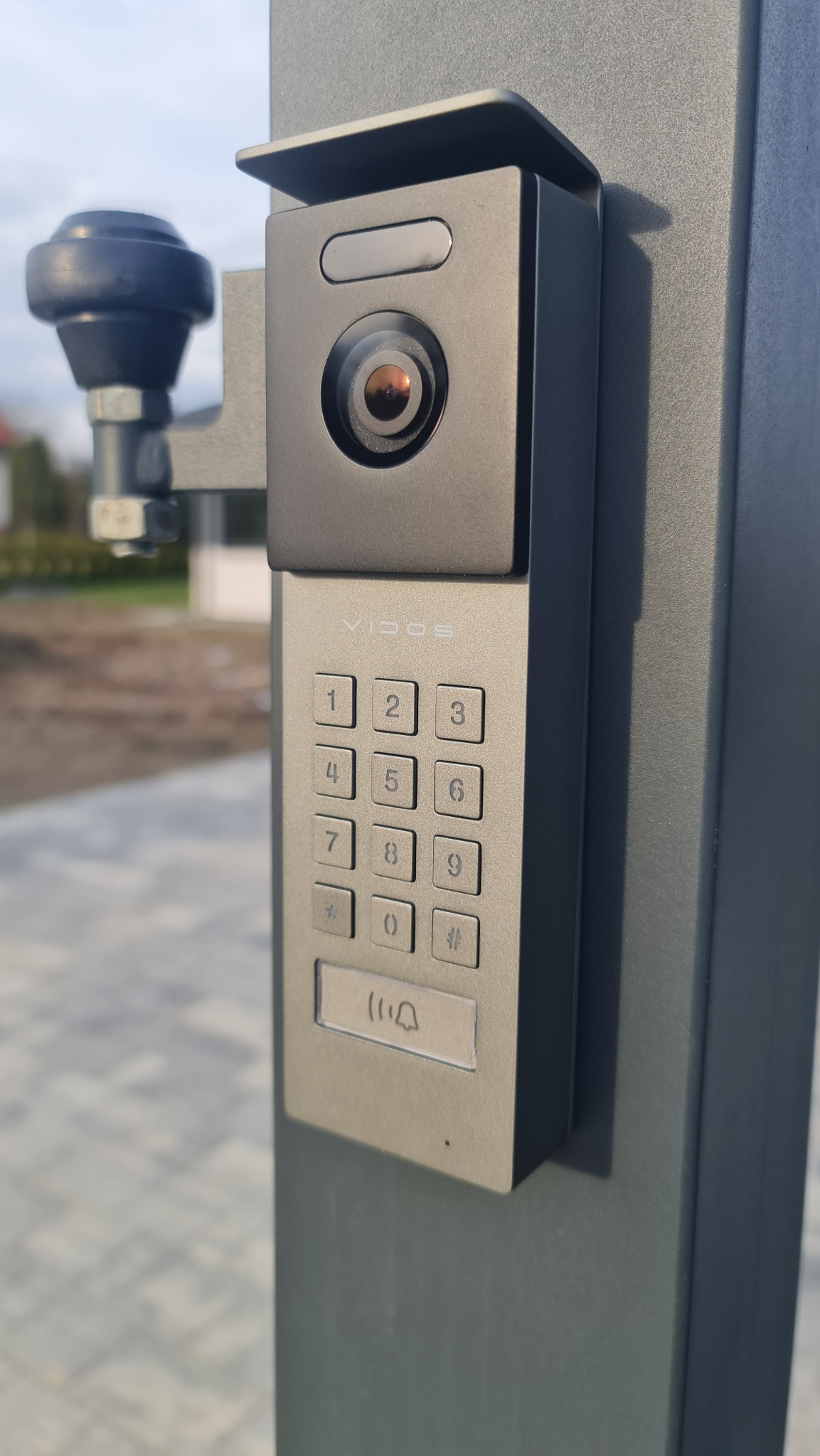 Szary wideofon Vidos z klawiaturą numeryczną i kamerą, zamontowany na metalowym słupku bramy wjazdowej, widok z bliska.