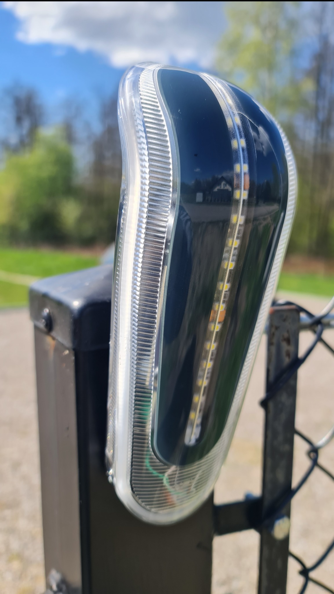 Lampa LED zamontowana na słupku bramy wjazdowej, widoczne diody i przezroczysta obudowa, tło z zielenią i błękitnym niebem.
