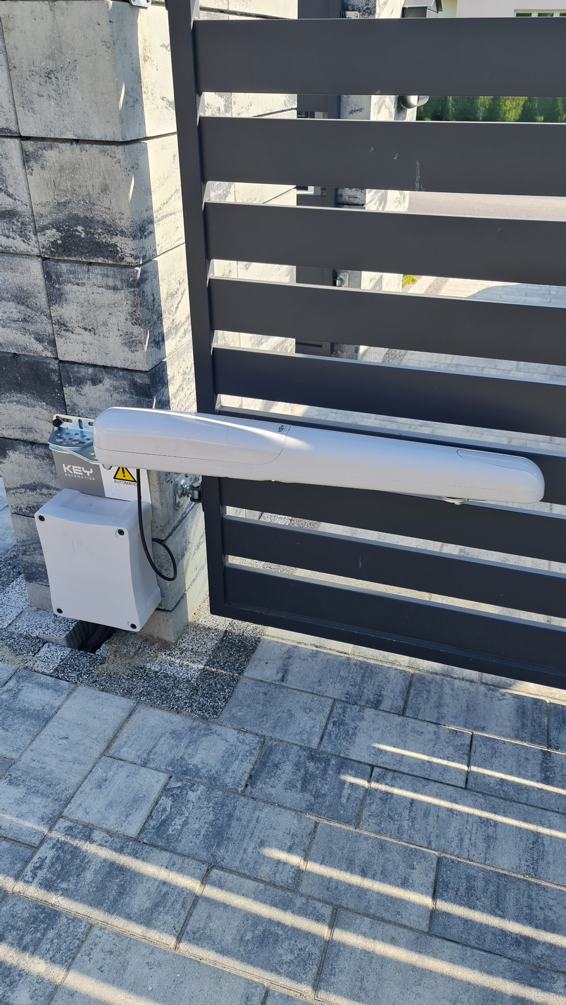 Szara brama posesyjna z zamontowanym napędem automatycznym KEY Automation, widoczna skrzynka sterująca z kablem i ostrzeżeniem o napięciu, kostka brukowa na podjeździe.