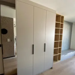 Jasnoszara szafa z trzema drzwiami i czarnymi uchwytami, z lustrem po lewej stronie i regałem z półkami po prawej, na tle jasnego parkietu.