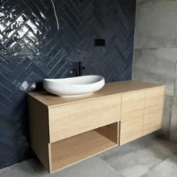 Nowoczesna łazienka: drewniana szafka z owalną umywalką na tle ciemnych, błyszczących płytek ułożonych w jodełkę. Minimalistyczny design, surowy beton na podłodze.