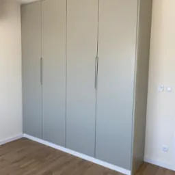 Szara, minimalistyczna szafa wnękowa z czterema drzwiami, z podłużnymi uchwytami, dopasowana do jasnego wnętrza z drewnianą podłogą.