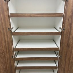 Bana Meble Rafał Korzeniowski - Otwarta szafa z półkami w kolorze drewna i białym wnętrzem. Widoczne zawiasy i metalowe prowadnice półek. Szafa z fornirem o wyraźnym rysunku słojów.