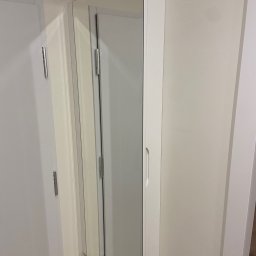 Bana Meble Rafał Korzeniowski - Biała szafka z lustrem, zamontowana na ścianie obok drzwi. Minimalistyczny design, widoczne zawiasy i uchwyt. Jasne wnętrze odbija się w lustrze.