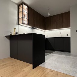 Nowoczesna kuchnia z czarną wyspą i drewnianymi szafkami. Podświetlane półki z kieliszkami. Kontrastujące podłogi: drewniana i szara płytka. Minimalistyczny design.