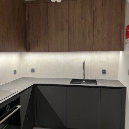 Nowoczesna kuchnia z ciemnymi szafkami dolnymi i drewnopodobnymi górnymi, oświetlona listwą LED. Minimalistyczny design z czarnym zlewem i baterią.