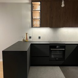 Nowoczesna kuchnia: czarne matowe fronty, drewniane szafki wiszące, szklana witryna z oświetleniem. Minimalistyczny design, szary blat i płytki. Wazon jako akcent kolorystyczny.