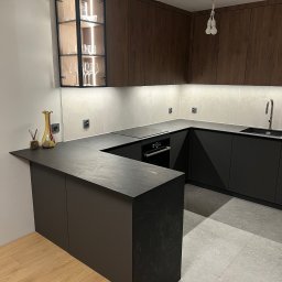 Nowoczesna kuchnia w kształcie litery U z ciemnymi frontami, blatem imitującym kamień i przeszkloną witryną z podświetleniem. Widoczne naczynia i dekoracje.