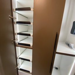 Bana Meble Rafał Korzeniowski - Otwarta szafa wnękowa z oświetleniem LED, brązowe fronty, wysuwane półki z czarnymi uchwytami. Widoczne zawiasy i fragment wnętrza drugiej części szafy.