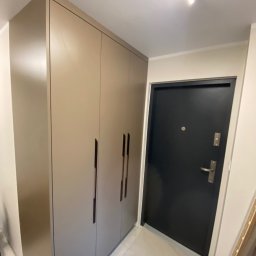 Bana Meble Rafał Korzeniowski - Beżowa szafa wnękowa z długimi, pionowymi uchwytami, umieszczona w przedpokoju obok czarnych drzwi wejściowych. Minimalistyczny design, jasne płytki podłogowe.